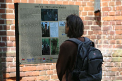 Mężczyzna stoi przed tablicą informacyjną dotyczącą kościoła i cmentarza w Grodźcu. W głębi fragment ściany z czerwonej cegły.