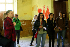 Grupa osób we wnętrzu zniszczonego budynku. Betonowa podłoga, zerwana boazeria i ściany upstrzone graffiti.