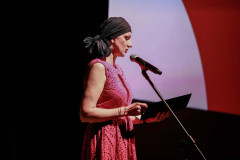 Sfotografowana z prawego profilu Marta Baran przy mikrofonie. Czyta z teczki. Ubrana w różową ażurową sukienkę. Na głowie chustka.