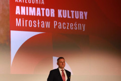 Na scenie Mirosław Pacześny w ciemnym garniturze, białej koszuli i różowym krawecie. Dłonie ukryte za plecami. W tle slajd z napisem: Kategoria Animator Kultury Mirosław Pacześny.