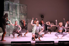 Tancerze Polskiego Teatru Tańca w trakcie występu. Kucają i wymachują prawymi dłońmi. Po lewej postać w kapturze z mikrofonem.