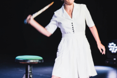 Aktorka wymachuje rakietą tenisowa trzymaną w prawej dłoni. Za nią stołek z zielonym siedziskiem.