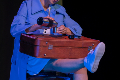 Aktorka siedzi na stołku ubrana w niebieski płaszcz. Na kolanach trzyma walizkę. Nalewa z termosu do kubka.