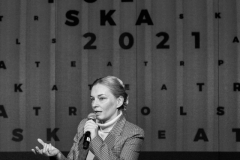 Agnieszka Przepiórska w fotelu. W lewej ręce trzyma mikrofon, prawą gestykuluje. W tle napis Teatr Polska 2021. Zjęcie czarno-białe