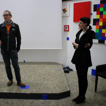 Na podwyższeniu stoi Robert Brzęcki. Lewa stopa na niebieskim kwadracie naklejonym na podłodze. Za nim biały ekran. Po prawej Rafaela Jesionowicz.