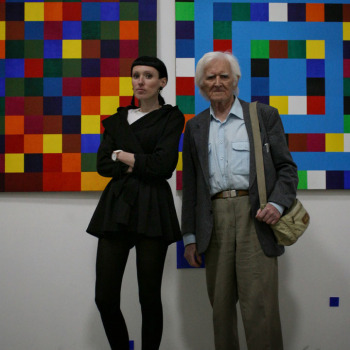 Rafaela Jesionowicz pozuje do zdjęcia ze starszym mężczyzną na tle prac, kompozycji złożonej z kolorowych kwadratów.