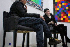 Siedzący w fotelach Robert Brzęcki i Rafaela Jesionowicz. Na ścianie wyeksponowane kolorowe prace. Zdjęcie z żabiej perspektywy.