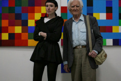 Rafaela Jesionowicz pozuje do zdjęcia ze starszym mężczyzną na tle prac, kompozycji złożonej z kolorowych kwadratów.