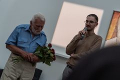 Po lewo Jan Sznajder trzyma bukiet czerwonych róż, po prawej Marcin Derda mówi do mikrofonu trzymanego w prawej dłoni.