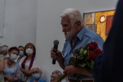 Jan Sznajder z bukietem czewronych róż. W prawej ręce trzyma mikrofon. W lewym dolnym rogu grupa uczestników wernisażu.