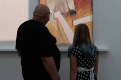 Stojący tyłem mężczyzna w czarnej koszulce i dziewczynka w sukience w białe grochy, przepasana białą szarfą oglądają jedną z prac.