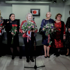 Grupa kobiet stoi przed małą sceną. Część z nich trzyma w dłoniach bukiety kwiatów. Przed nimi mikrofon na statywie. 