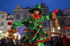 Artystka w zielonym świątecznym stroju i zielonym kapleuszu uśmiecha się do obiektywu. Rozkłada w bok ręce. W tle publiczność i kamienica przy placu.