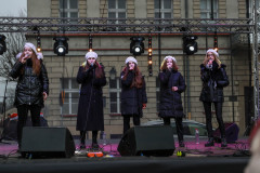 Pięć wokalistek na scenie. Ubrane w ciemne płaszcze lub kurtki i jasne berety.