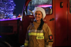 Uśmiechnięta kobieta w uniformie strażackim pozuje do zdjęcia. Za nią fragment czerwonego wozu.