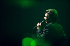 Wojciech Myrczek śpiewa do trzymanego w lewej dłoni mikrofonu.  Na zdjęciu widać zielone światła.