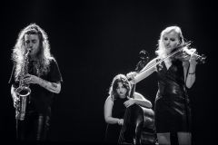 Po lewej: Monika Muc gra na saksofonie, po prawej Dominika Rusinowska gra na skrzypcach, za nimi kontrabasistka Kamila Drabek. Zdjęcie czarno-białe.
