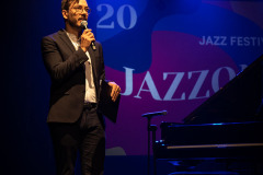 Ubrany w garnitur Andrzej Zieliński trzyma mikrofon w prawej dłoni. Po prawo fortepian. W tle wyświetlony slajd Jazzonaliów. Rzecz się ma na scenie CKiS-DK Oskard.