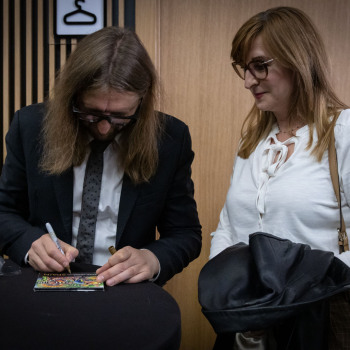 Leszek Możdżer podpisuje płytę. Po prawej oczekująca na autograf kobieta w białej bluzce trzyma czarną kurtke.