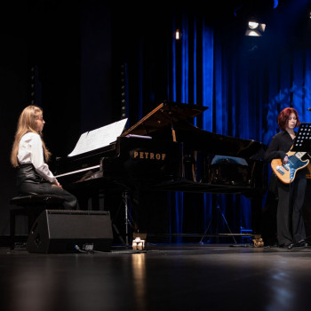 Po lewej przy fortepianie Petrof siedzi dziewczyna z rozpuszczonymi włosami, po prawej gitarzystka przy pulpicie. Na pierwszym planie w lewym dolnym rogu żarówka.