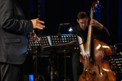 Sfotografowany z prawego profilu Krzysztof Pydyński przy pulpicie dyrygenckim. W głębi kadru Szymon Wieczorkiewicz gra na kontrabasie.