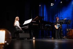 Po lewej przy fortepianie Petrof siedzi dziewczyna z rozpuszczonymi włosami, po prawej gitarzystka przy pulpicie. Na pierwszym planie w lewym dolnym rogu żarówka.