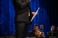 Stojący bokiem Paweł Zieliński. W lewej dłoni trzyma mikrofon, w prawej podkładkę. W głębi kadru Big Band.