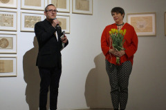 Joanna Imielska (po prawej) ubrana w czerwoną koszulę i spodnie we wzorek trzyma bukiet żółtych tulipanów. Po lewej Robert Brzęcki w czarnej marynarce mówi do mikrofonu. Oboje stoją na podwyższeniu. Za nimi prace wyeksponowane na jasnej ścianie.