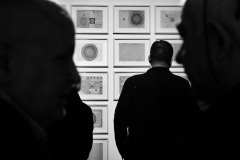 Po bokach zdjęcia ciemne profile rozmawiających mężczyzn. Pośrodku stojący tyłem do obiektywu mężczyzna ogląda prace. Zdjęcie czarno-białe.
