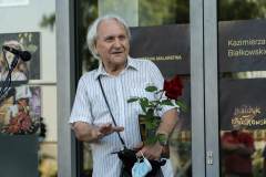 Uśmiechnięty Kazimierz Białkowski trzyma czerwoną różę. Gestykuluje prawą ręką.