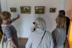 Korytarz budynku przy Okólnej. Goście wernisażu wraz z artystą oglądają prace. Kobieta po lewo wskazuje lewą ręką jeden z obrazów.
