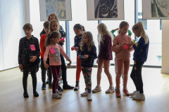 Grupa dzieci w sali multifunkcyjnej CKiS-DK Oskard. Mają poprzypinane do koszulek karteczki lub trzymają je w dłoniach. W tle przeszklona ściana i wiszące prace.