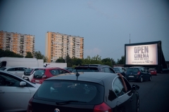 W prawym górnym rogu dużych rozmiarów ekran kina samochodowego. Wyświetlany jest na nim napis "Open Cinema. Mobilne kino Charlie". Widok z miejsca parkingowego. W tle po lewej stronie dwa wieżowce.