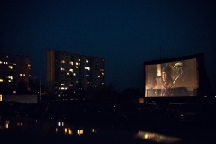 Ekran kina samochodowego widziany z miejsca parkingowego.  W tle po lewej stronie dwa wieżowce. Zapada zmrok.