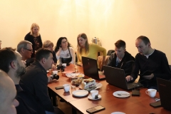 Członkowie Konińskiego Klubu Fotograficznego podczas spotkania w budynku Centrum Kultury i Sztuki przy ulicy Okólnej.  Siedzą po obu stronach długiego stołu, który zastawiony jest laptopami, słodkim poczęstunkiem, białymi talerzykami i filiżankami.