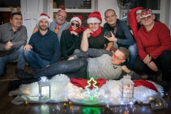 Grupa uśmiechniętych mężczyzn kuca na podłodze. Na głowach świąteczne ozdoby lub mikołajkowe czapki. Jeden z mężczyzn leży. Przed nimi świąteczne lampiony. W tle okna.