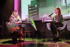Anna Pilarska i Monika Dolata z mikrofonem w trakcie rozmowy. Siedzą w szarych fotelach. Przed nimi stoliki z karafkami i szklankami wody.