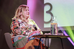 Anna Pilarska siedzi w fotelu. Ubrana w kolorową spódnicę i koszulę z portretami Fridy Kahlo. Przed nią stolik, a na nim karafka i szklanka. W tle fragment slajdu. Wyświetlona cyfra 3.