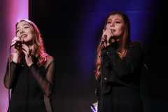 W trakcie występu. Dwie dziewczyny w czerni śpiewają trzymając w dłoniach mikrofony.