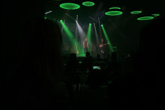 Ciemna sala. Sylweetki publiczności. W głębi kadru scena, a na niej artyści. Zielone światła reflektorów.