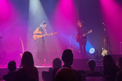 Sylwetki siedzących widzów odwrócnych plecami do obiektywu. Na scenie Misia Furtak i mężczyzna. Obydwoej mają gitary. Różowe światła reflektorów.