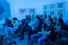 Publiczność siedzi na czarnych plastikowych krzesełkach ustawionych w rzędy. Zdjęcie utrzymane w błękitnej tonacji.