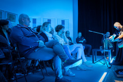 Publiczność siedząca na krzesełkach. Po prawej jeden z członków zespołu. Zdjęcie zrobione z żabiej perspektywy, utrzymane w błękitnej tonacji.