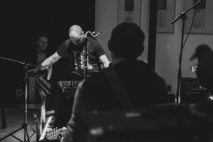 Zespół w trakcie koncertu. Gitarzysta odwrócony plecami do obiektywu. Przodem z pochyloną głową Krzysztof Matysiak. Zdjęcie czarno-białe.