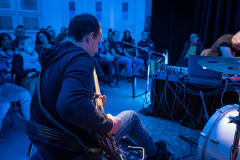 Sfotografowany tyłem gitarzysta siedzi na krześle i gra. W głębi kadru publiczność.