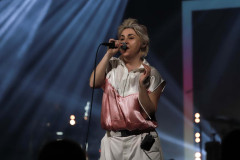 Maria Peszek na scenie. Śpiewa do mikrofonu trzymanego w prawej dłoni. Ubrana w białe spodnie i biało-różową koszulkę. Zdjęcie w planie amerykańskim.