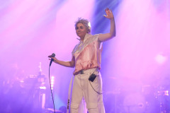 Maria Peszek na scenie. Ubrana w białe spodnie i biało-różową koszulkę. Macha rękoma. W prawej trzyma mikrofon.