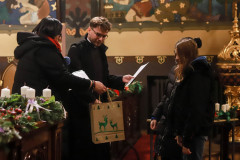Renata Rudowicz podaje papierową torbę z rysunkiem dwóch zielnych jeleni. Ksiądz trzyma kartkę. Przed nimi dwie uczestniczki konkursu sfotografowane z lewego profilu.
