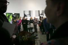 Uczestnicy wystawy "THE PRESENT – Państwowe Liceum Sztuk Plastycznych w Kościelcu" zgromadzeni w galerii CKiS "Wieża Ciśnień". Na pierwszym planie dwie zamglone sylwetki mężczyzn.