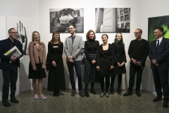 Twórcy prac wystawianych w galerii CKiS "Wieża Ciśnień" podczas "THE PRESENT – Państwowe Liceum Sztuk Plastycznych w Kościelcu". Stoją rzędem pod białą ścianą, na której zawieszono fotografie.